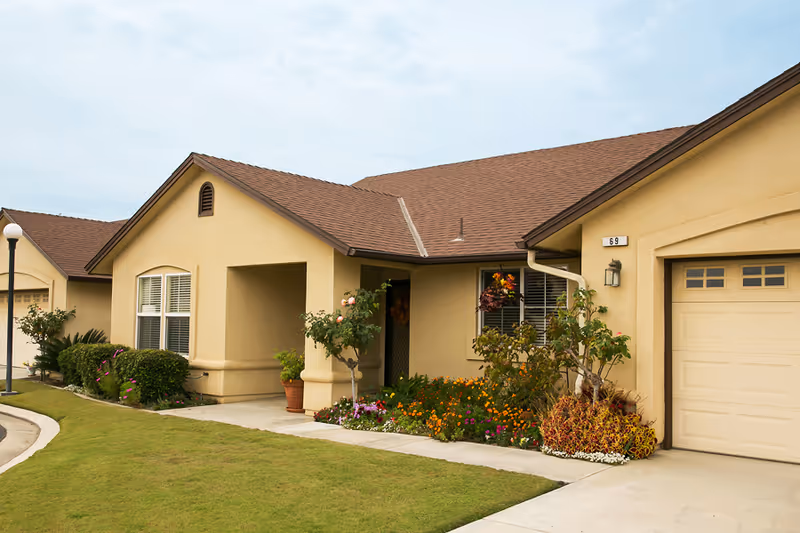Single-story beige retirement community cottage front with attached garage, lawn, and colorful flower beds.