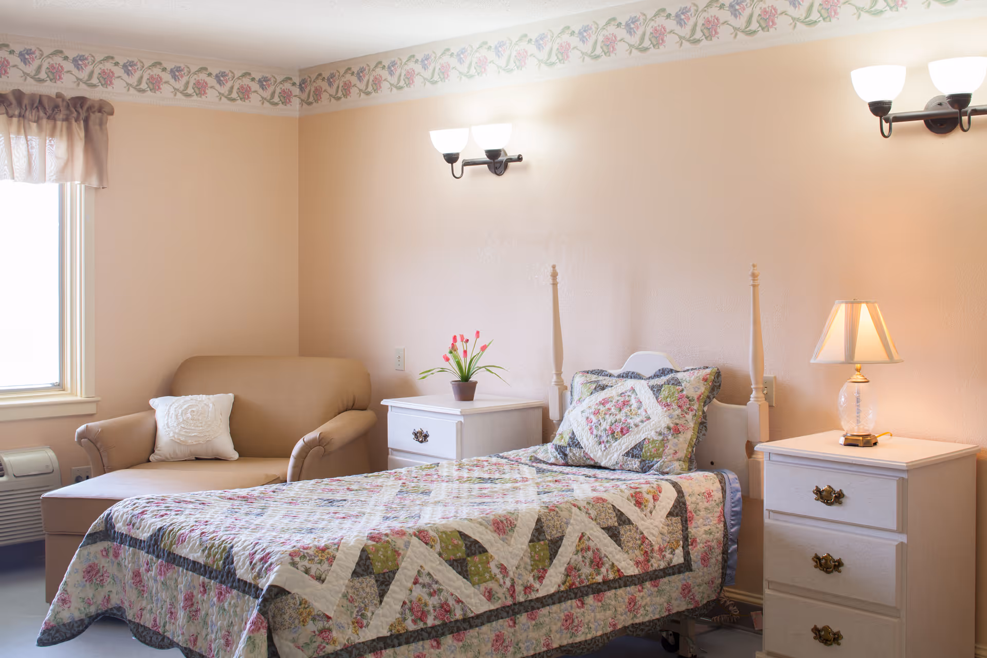 A cozy single bedroom with a floral quilted bed, an armchair by the window, nightstands with a lamp and flowers, and soft pastel walls.