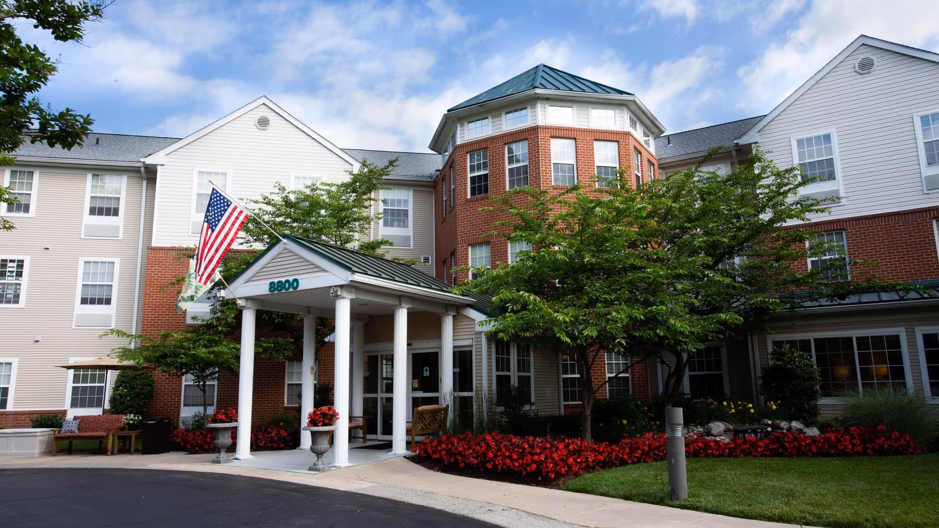 Front entrance of a brick-and-siding senior living building with a covered porte-cochere, American flag, trees, and red flower beds.