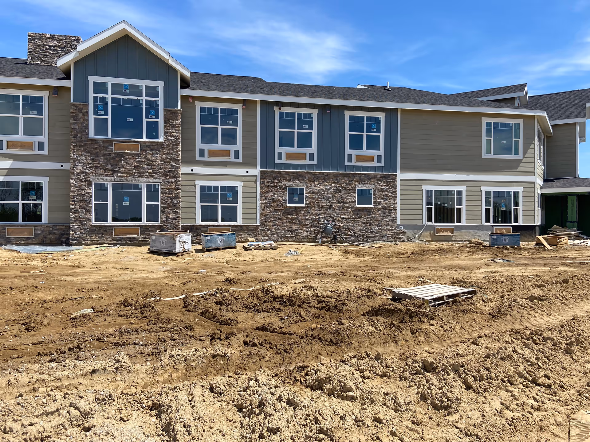 Exterior view of a two-story building under construction with stone and siding facade, multiple windows, and a dirt area in front of the building.