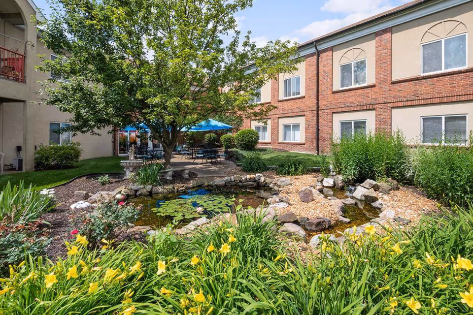 Outdoor garden area at Richmond Terrace featuring a small pond with lily pads surrounded by rocks and various plants, including yellow flowers. There is a tree providing shade over a patio area with tables, chairs, and blue umbrellas. The background shows a two-story brick and beige building with multiple windows.