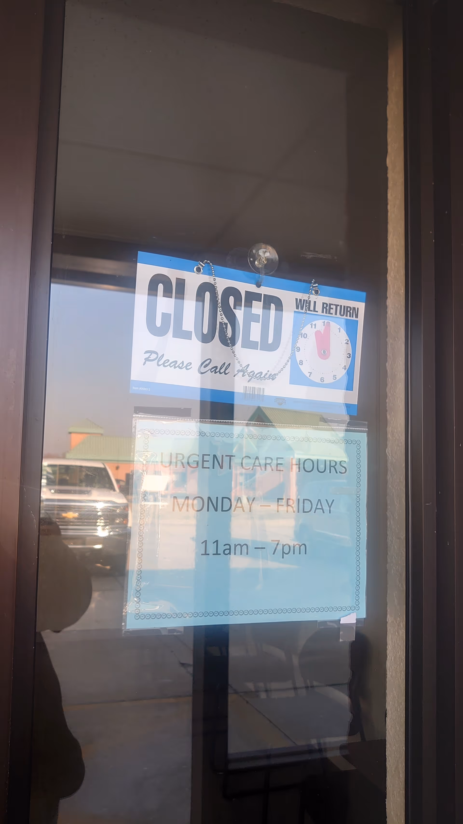 Glass entrance door with closed sign and urgent care hours posted, reflecting outside scene.