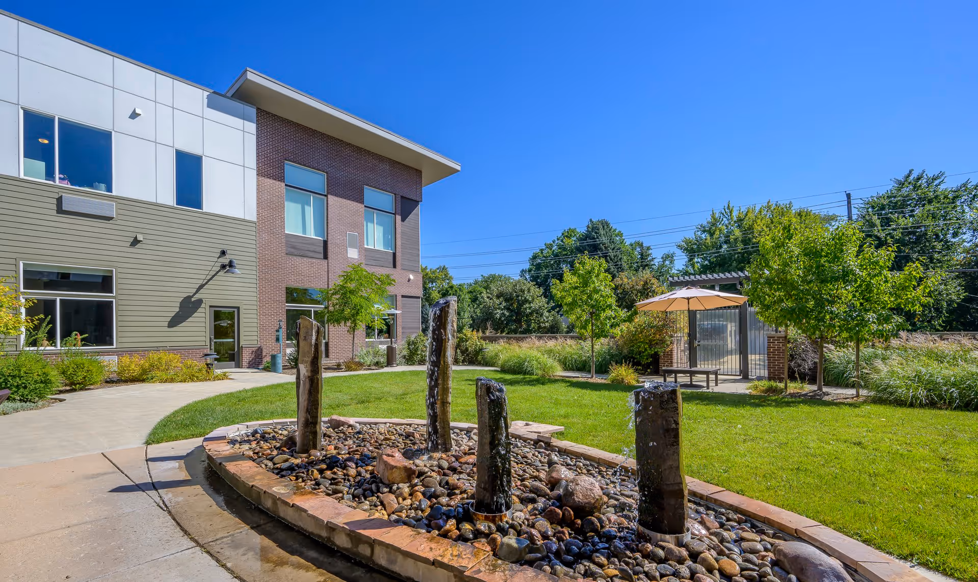 Outdoor garden area at Rosemark At Mayfair Park featuring a modern water fountain with four vertical stone pillars surrounded by rocks, a green lawn, small trees, a paved walkway, and a building with large windows under a clear blue sky.