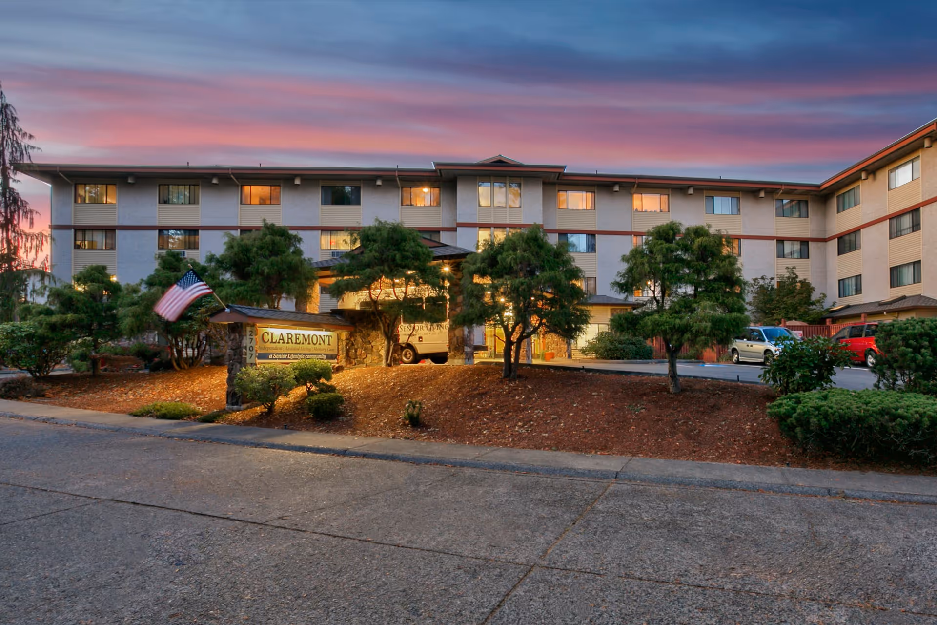 Exterior front view of a multi-story senior living building at dusk with a lit entrance, landscaped grounds, parked cars, and a sign reading 'Claremont'.