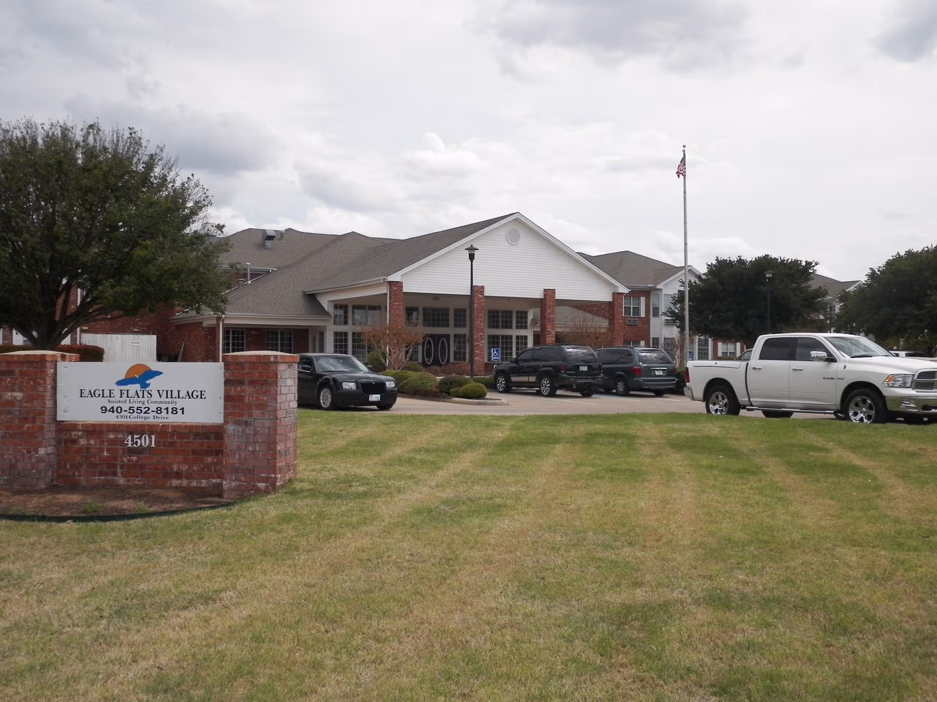 Exterior view of Eagle Flats Village assisted living community building with a brick sign in the foreground displaying the facility name, phone number, and address. Several cars are parked in front of the building, and an American flag is flying on a flagpole near the entrance.