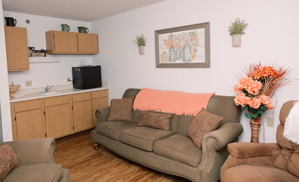 A cozy living area with a green upholstered sofa adorned with patterned pillows and a coral throw blanket. To the right is a brown recliner chair with a white throw. Behind the sofa is a wall decorated with a framed floral painting and two small hanging planters. On the left side of the room is a small kitchenette with wooden cabinets, a black mini fridge, a sink, and some decorative items on the countertop and shelves. The floor is wooden.