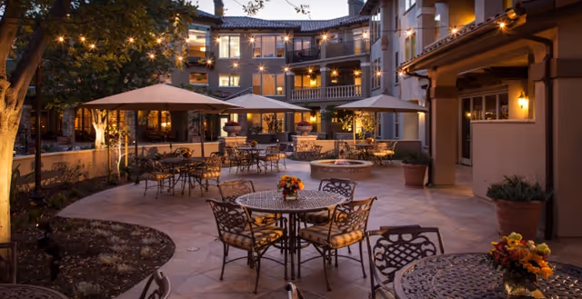 Outdoor courtyard area at dusk with multiple round metal tables and chairs, some tables decorated with flower arrangements. Large umbrellas provide shade, and string lights are hung above. The courtyard is surrounded by a multi-story building with balconies and warm lighting from windows and wall sconces.