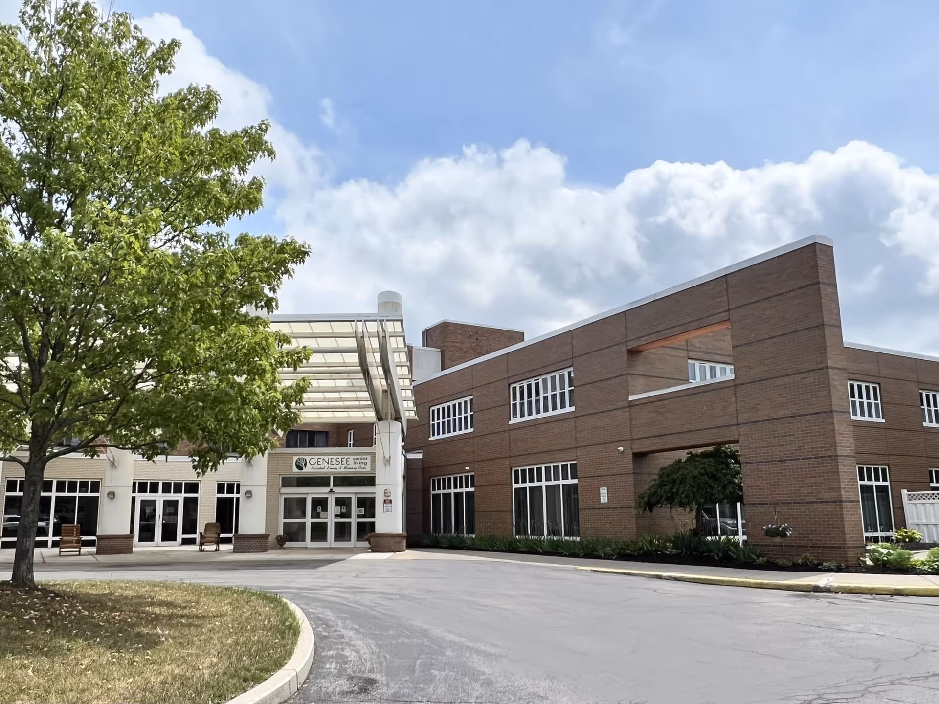 Exterior view of Genesee Senior Living facility showing a two-story brick building with multiple windows, a covered entrance, a tree on the left, and a driveway in front under a partly cloudy sky.