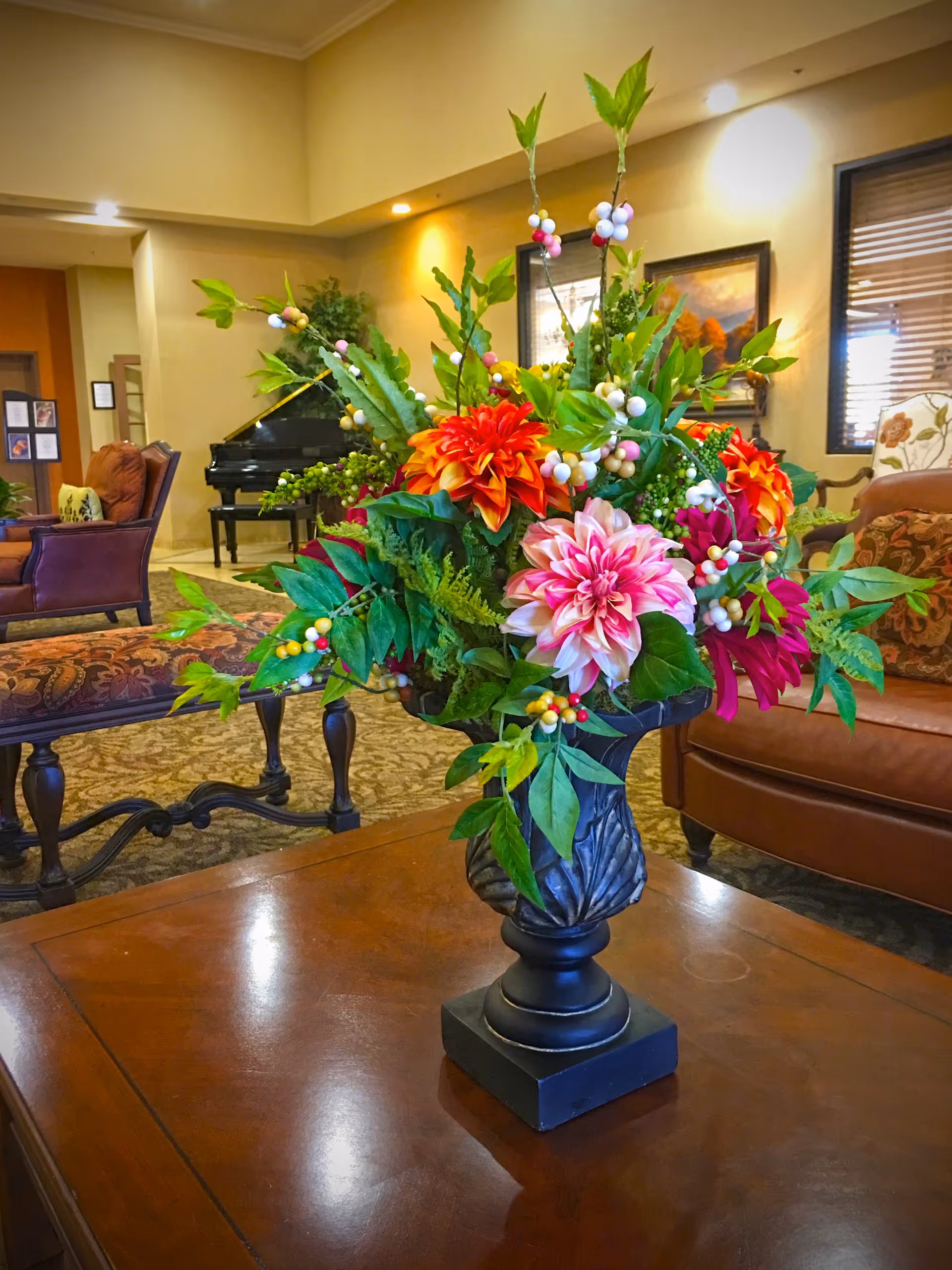 A decorative floral arrangement with vibrant orange, pink, and red flowers along with green leaves and white berries in a black vase sits on a wooden table in a warmly lit living room. The background features upholstered chairs, a piano, framed artwork, and a window with blinds.