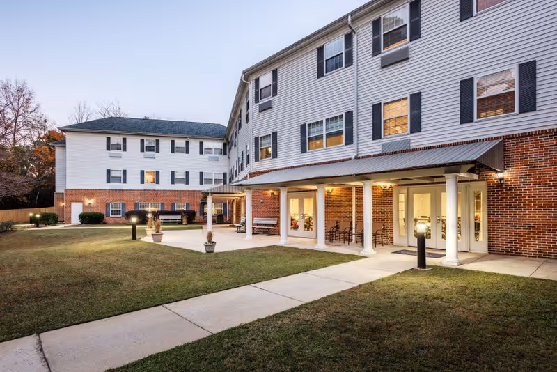 Exterior view of Oakleaf Village of Lexington, showing a multi-story building with white siding and red brick accents. The building has several windows with black shutters and a covered entrance supported by white columns. There is a paved walkway and a grassy lawn in front, with outdoor lighting and some potted plants near the entrance.