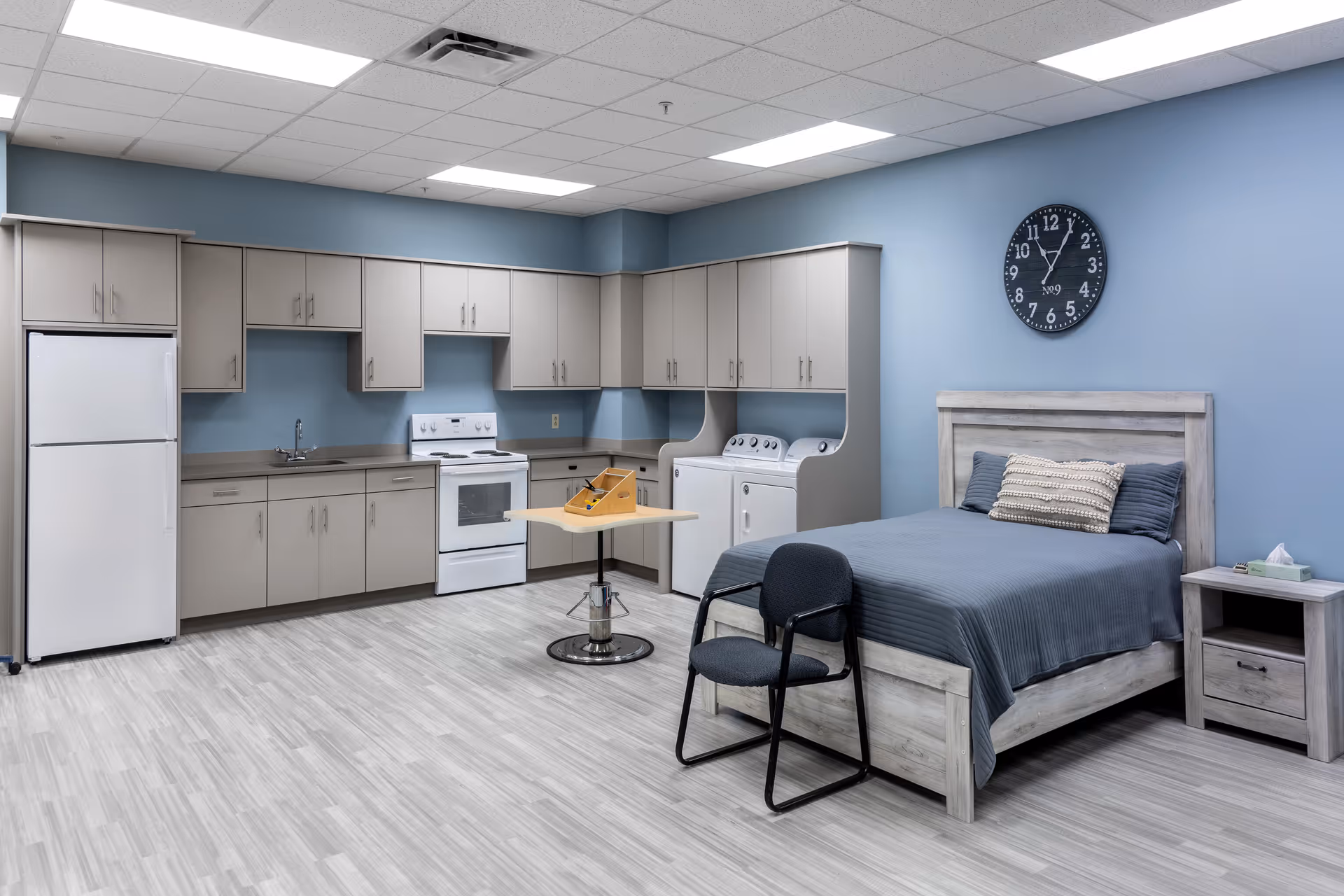 A spacious room with a kitchenette featuring a refrigerator, stove, sink, and multiple cabinets. Next to the kitchenette are a washer and dryer set. The room also contains a bed with blue bedding, a nightstand with tissues and a book, a chair, and a small table with a wooden game on it. The walls are painted light blue, and a black wall clock shows the time as 12:00.