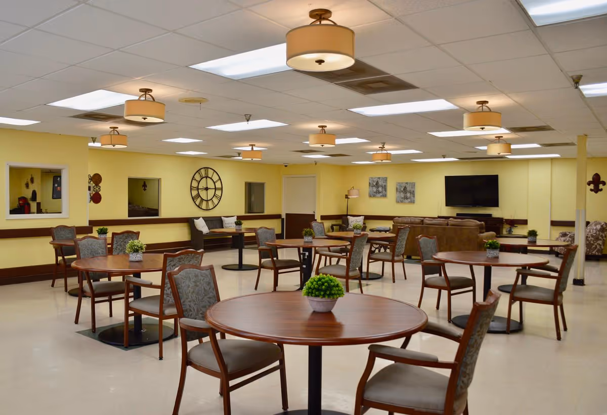 A spacious community room with several round wooden tables and cushioned chairs arranged neatly. The walls are painted yellow with dark brown trim. There are decorative plants on each table, a large wall clock, and a flat-screen TV mounted on the far wall. Comfortable sofas and armchairs are placed near the TV, and ceiling lights illuminate the room.