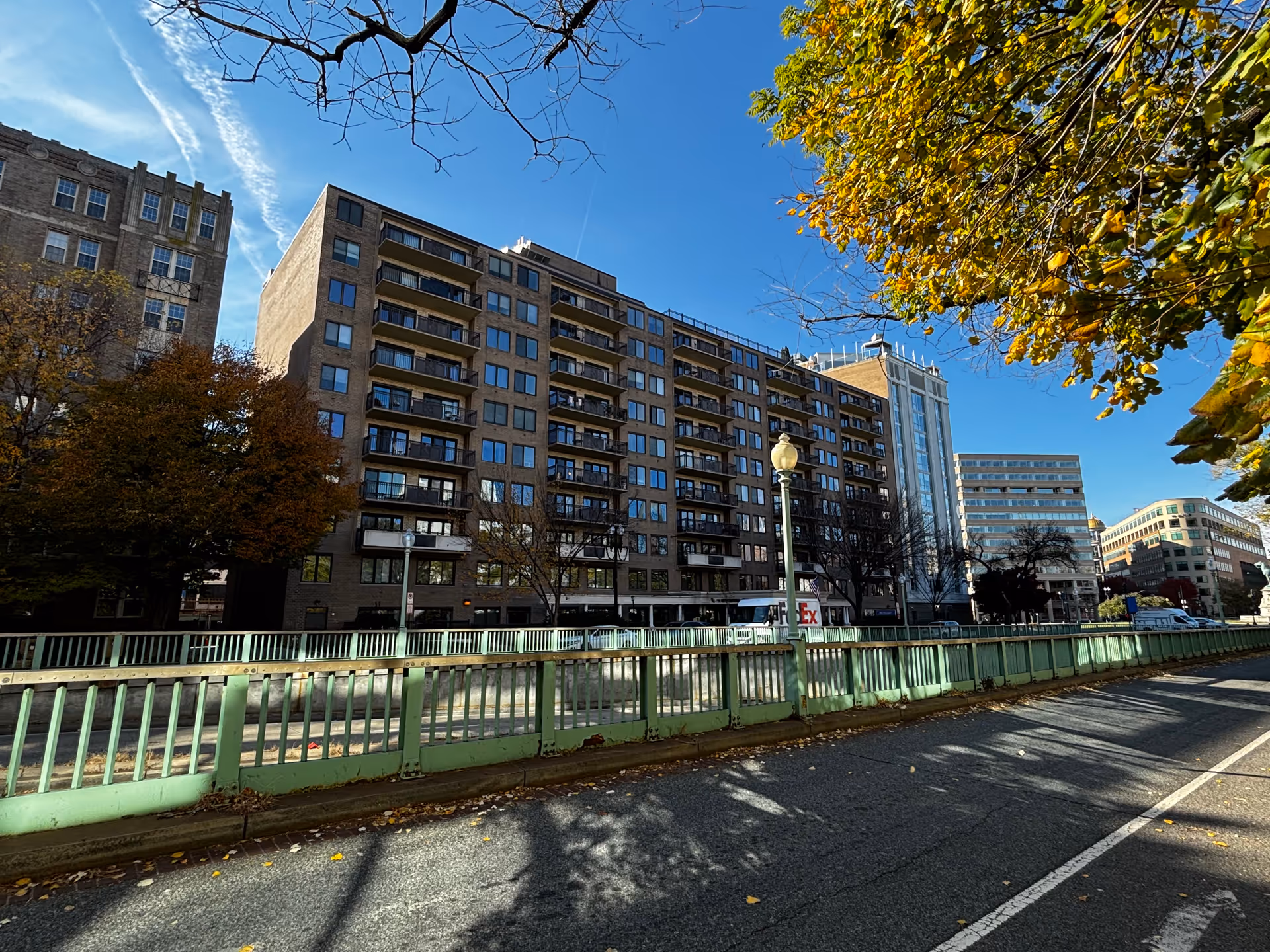 Street-level view of a multi-story brick apartment building with balconies, trees, and a green railing along the road under a clear blue sky.