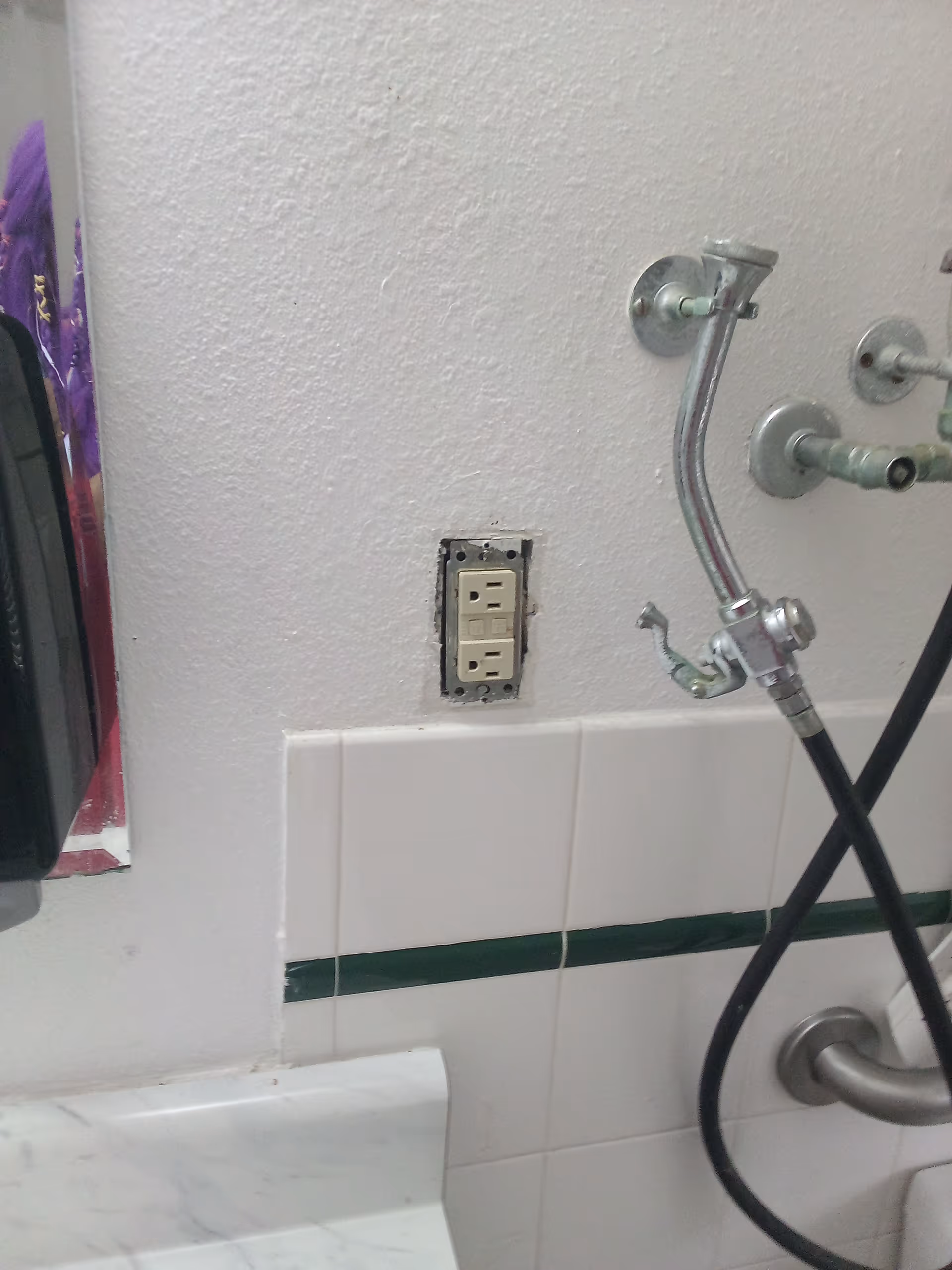 Bathroom wall with exposed outlet and shower hoses beside tiled surface