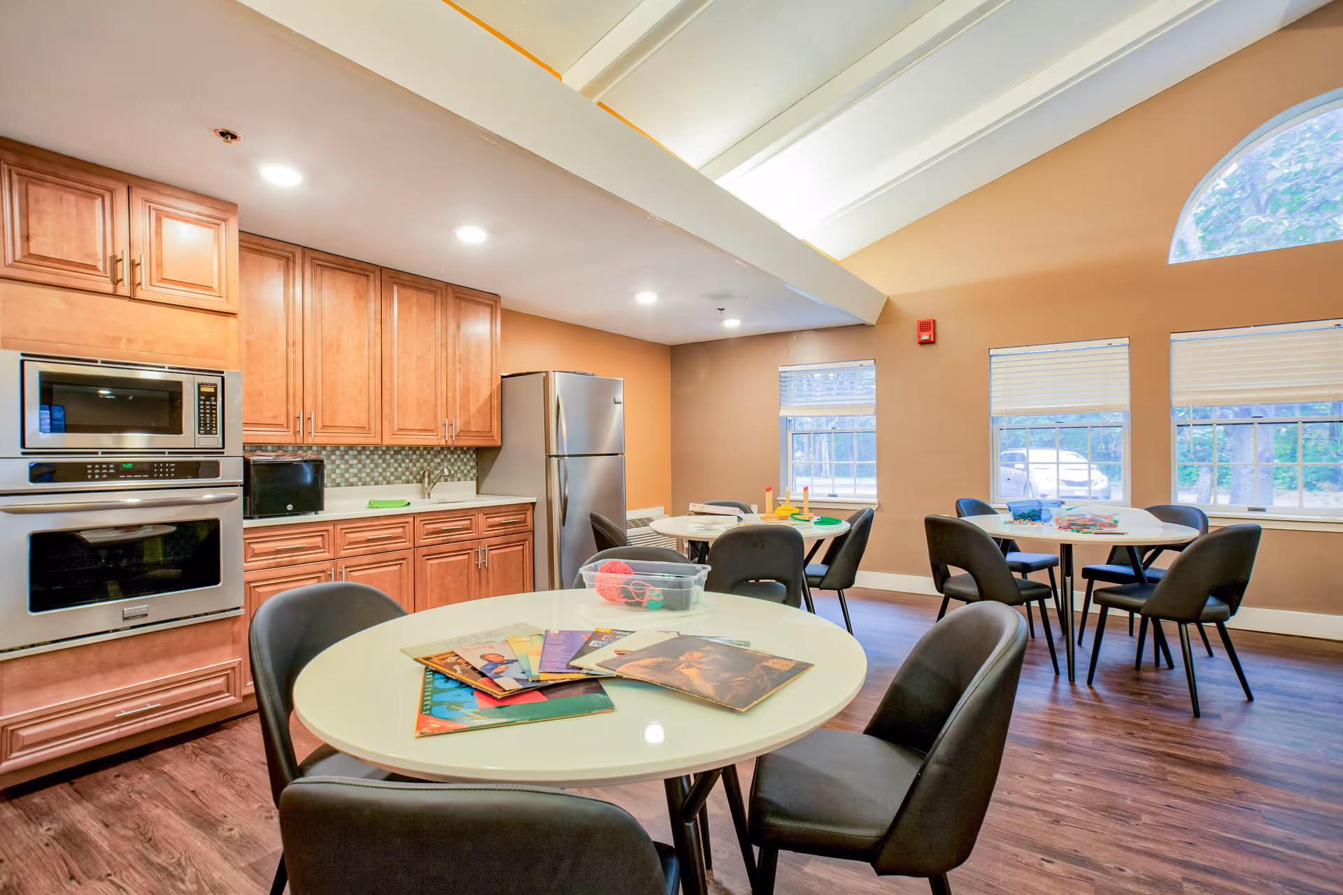 Bright communal kitchen and dining area with round tables, chairs, and wooden cabinets.