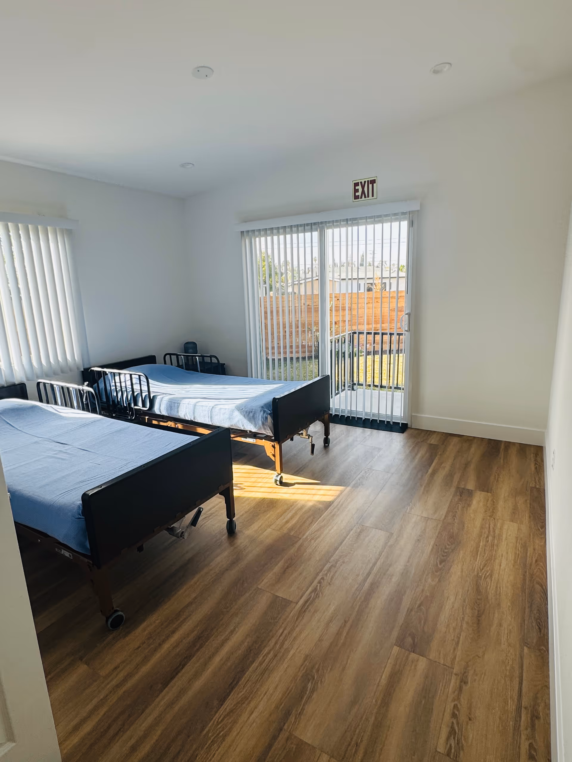 Two hospital-style beds in a bright, minimally furnished bedroom with an exit door.