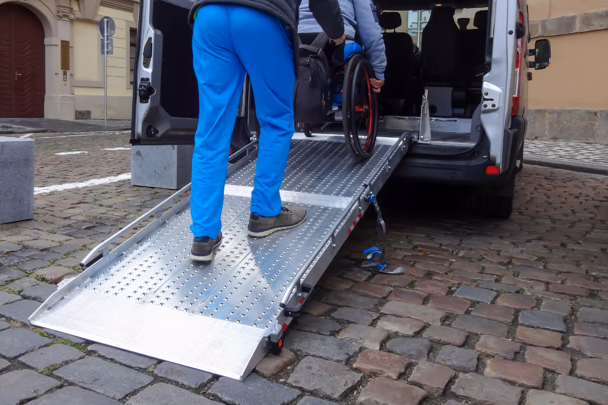 Caregiver helps wheelchair user board a van using a ramp outdoors.