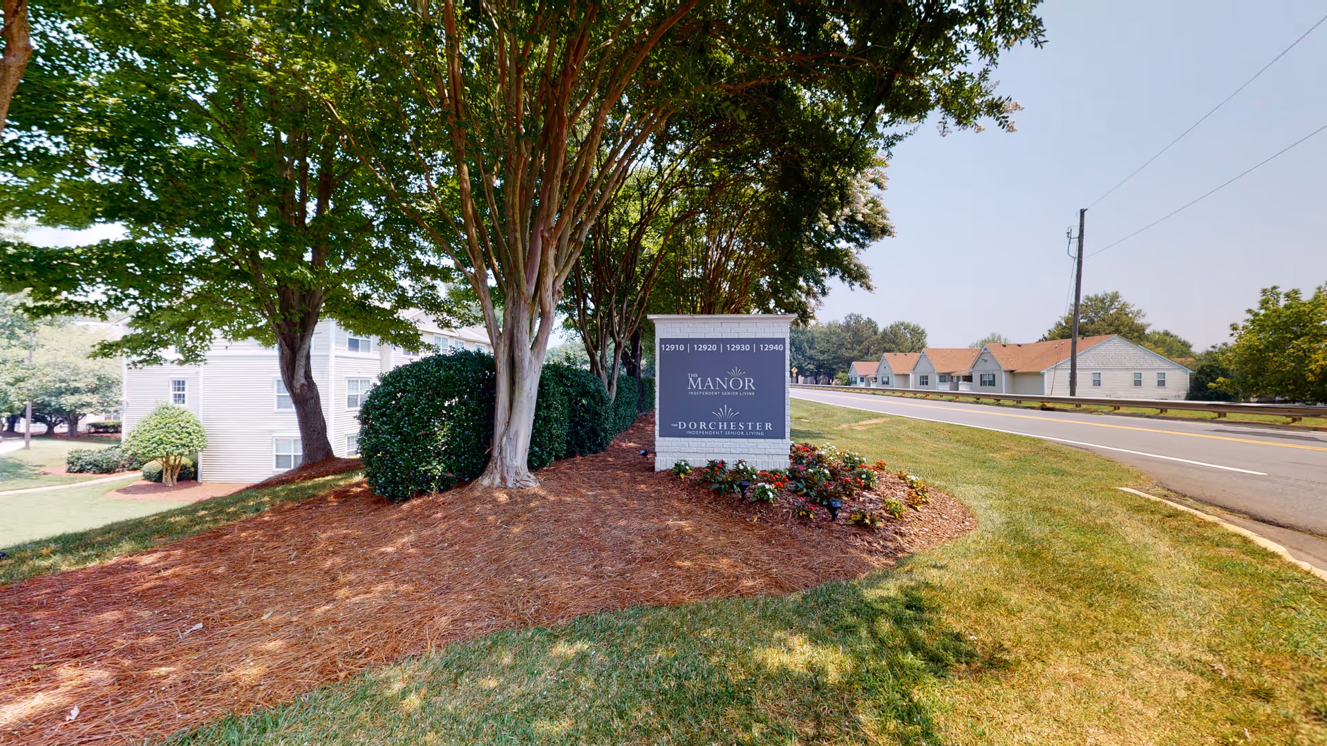Landscaped entrance sign for The Dorchester Manor beneath trees by a roadway with nearby buildings.