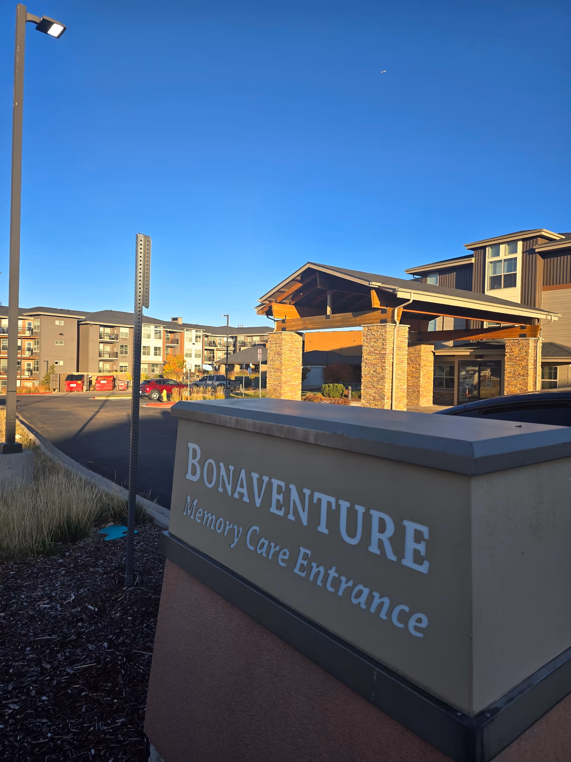 Entrance sign reading 'Bonaventure Memory Care Entrance' in front of a multi-story senior living building with a covered porte-cochere under a clear blue sky.
