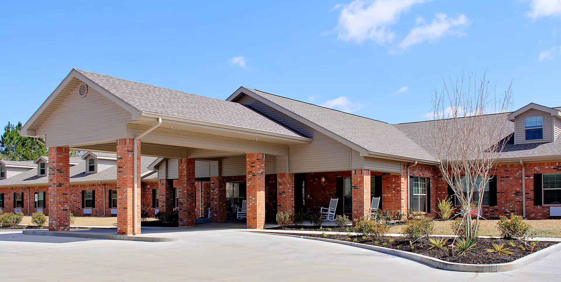 Exterior view of Gulfport Care Center showing a single-story brick building with a covered entrance supported by brick columns, a driveway, and landscaped areas with small plants and trees under a clear blue sky.