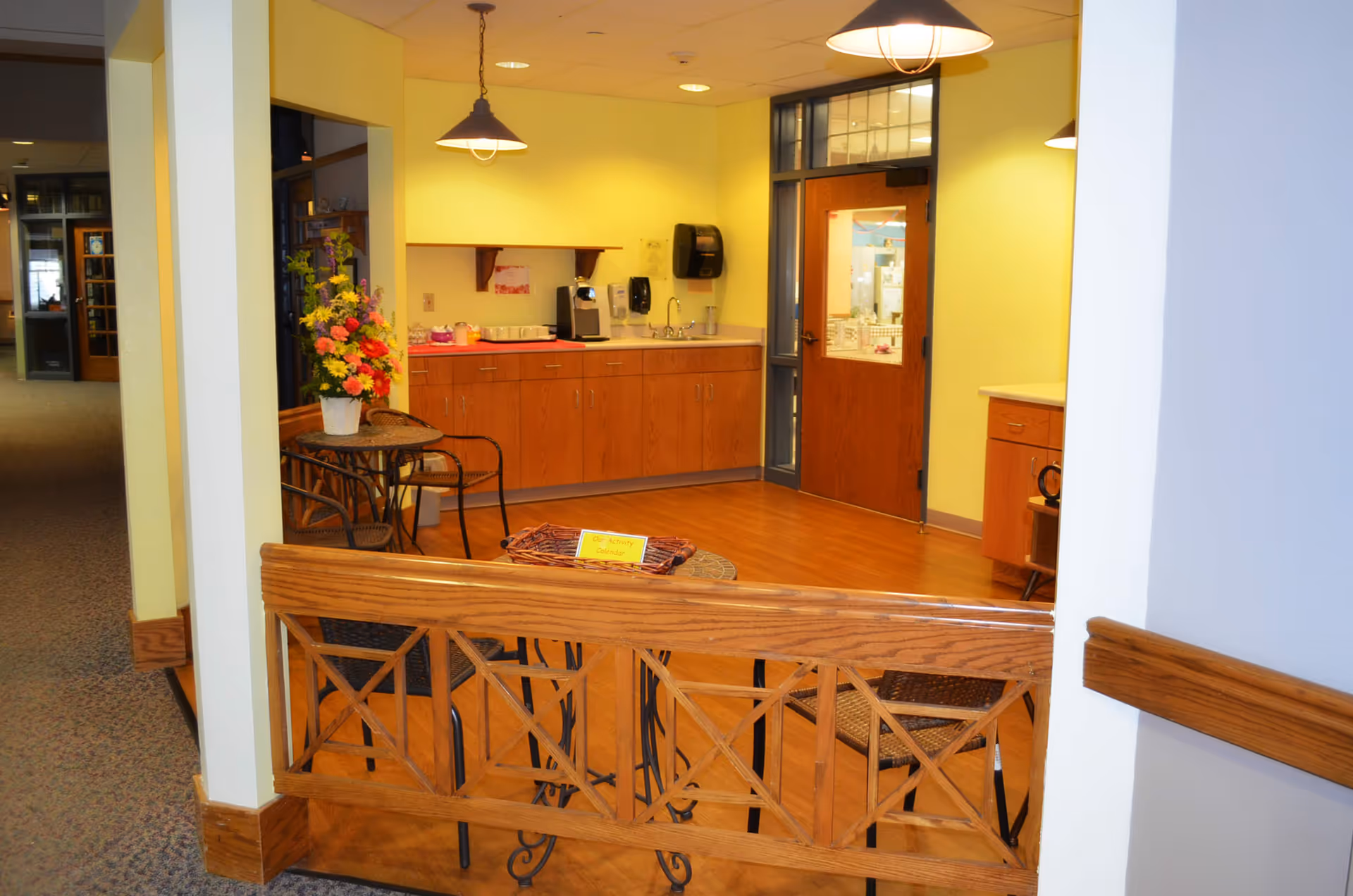 Small common area with a wooden railing, a round table and chairs, a floral arrangement, and a kitchenette with cabinets and pendant lights.