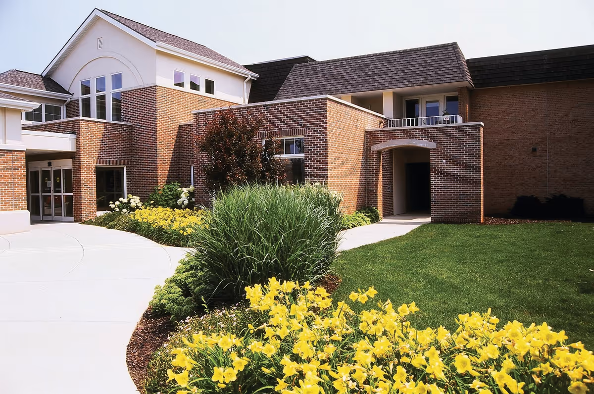 Exterior view of Porter Hills Village building featuring brick walls, large windows, a walkway, and landscaped garden with yellow flowers and green shrubs.