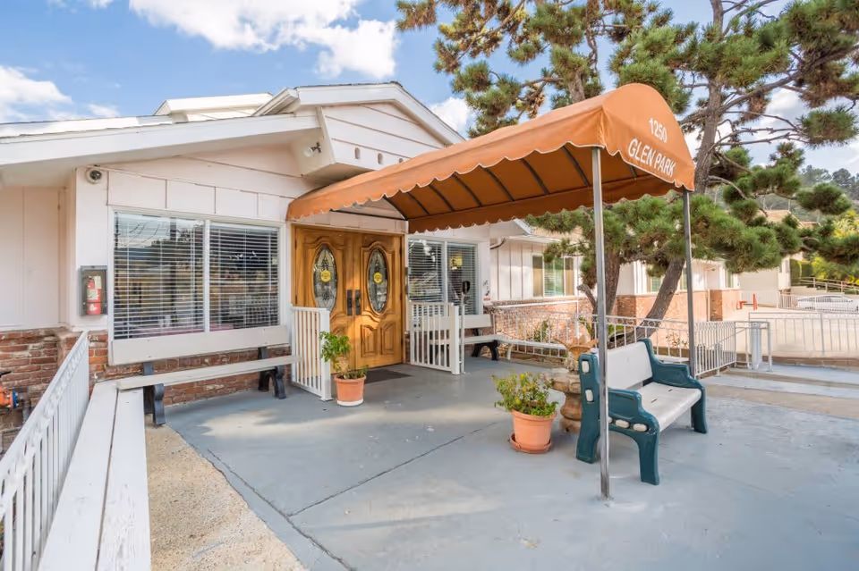 Entrance of Glen Park at Glendale - Mariposa facility with double wooden doors under an orange awning that reads '1250 Glen Park'. There are benches and potted plants on the concrete patio area, with trees and a clear sky in the background.