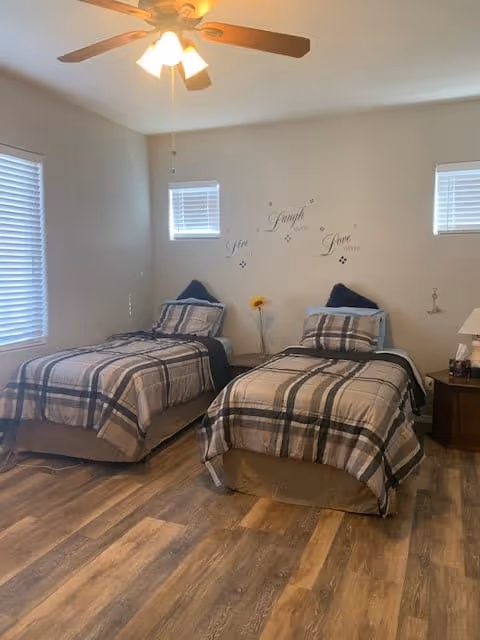 A bedroom with two single beds side by side, each with plaid bedding in shades of gray, beige, and black. There are two small windows on the wall behind the beds, and a ceiling fan with lights above. A small nightstand with a lamp and some items is positioned to the right of the beds. The wall above the beds has decorative text that reads 'Live Laugh Love' with small decorative elements around it. The floor is wood with a natural finish.