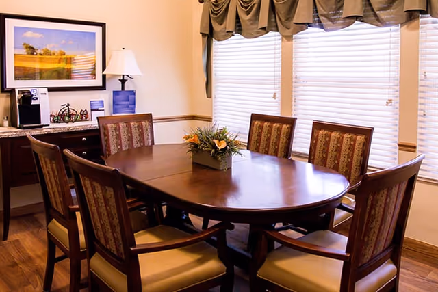 A dining room with a polished wooden oval table surrounded by six upholstered chairs. A floral centerpiece is placed on the table. The room has large windows with blinds and valances, a framed landscape picture on the wall, a table lamp, and a coffee maker on a side cabinet.