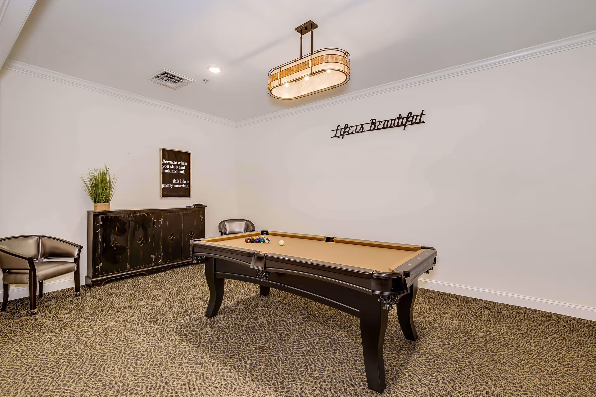 A recreational room with a pool table in the center, two chairs against the wall, a dark wooden cabinet with a potted plant on top, and wall decorations including a sign that reads 'Because when you stop and look around, this life is pretty amazing' and a metal wall art piece that says 'Life is Beautiful'.
