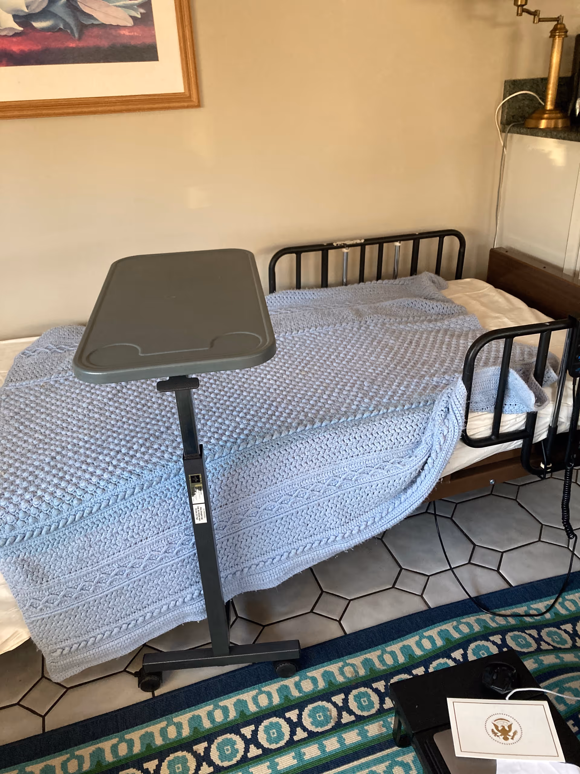 Hospital-style bed with blue knitted blanket and overbed table in a bedroom.