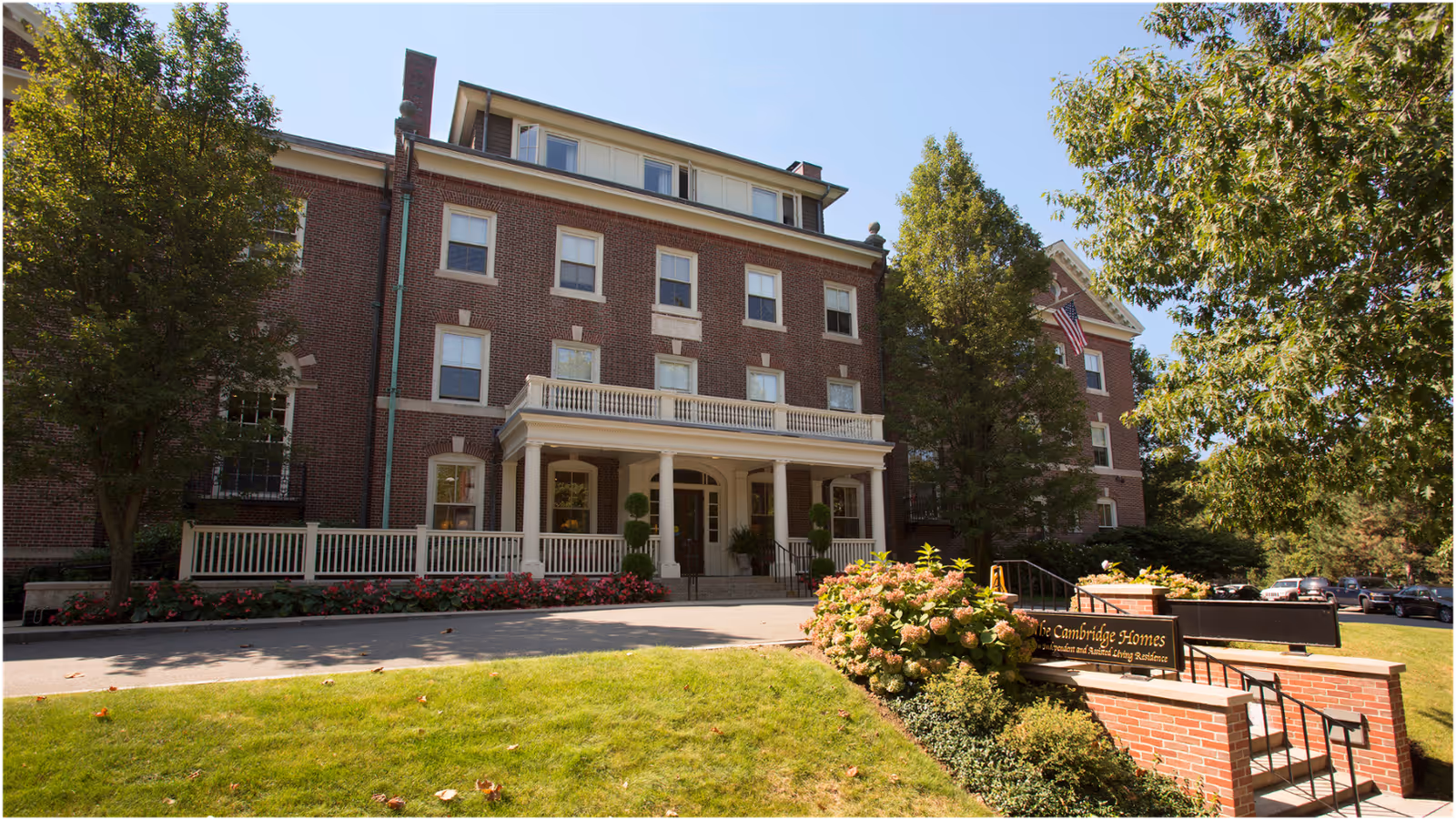Exterior view of a large brick building with white trim and multiple windows, surrounded by trees and greenery. There is a porch with columns at the entrance and a sign in front that reads 'The Cambridge Homes'. The sky is clear and blue.