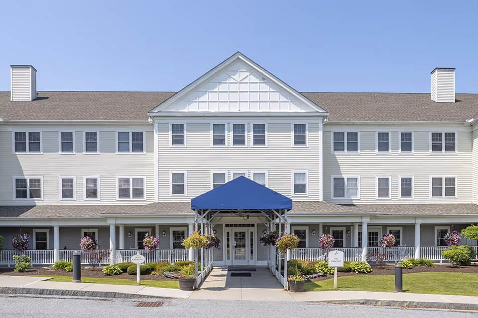 Front facade of a three-story senior living building with a blue entrance awning and hanging flower baskets.