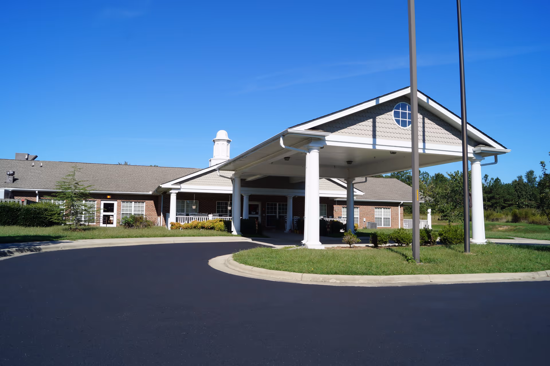 Front exterior view of a single-story brick building with a covered driveway supported by white columns, surrounded by green grass and bushes under a clear blue sky.