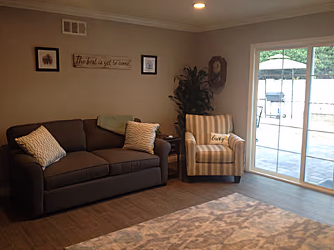 A cozy living room with a dark gray sofa adorned with two patterned pillows, a striped armchair with a pillow labeled 'Lucky', a small side table with a plant, and a sliding glass door leading to an outdoor patio with a table and umbrella. The walls are decorated with framed pictures and a sign that reads 'The best is yet to come.'