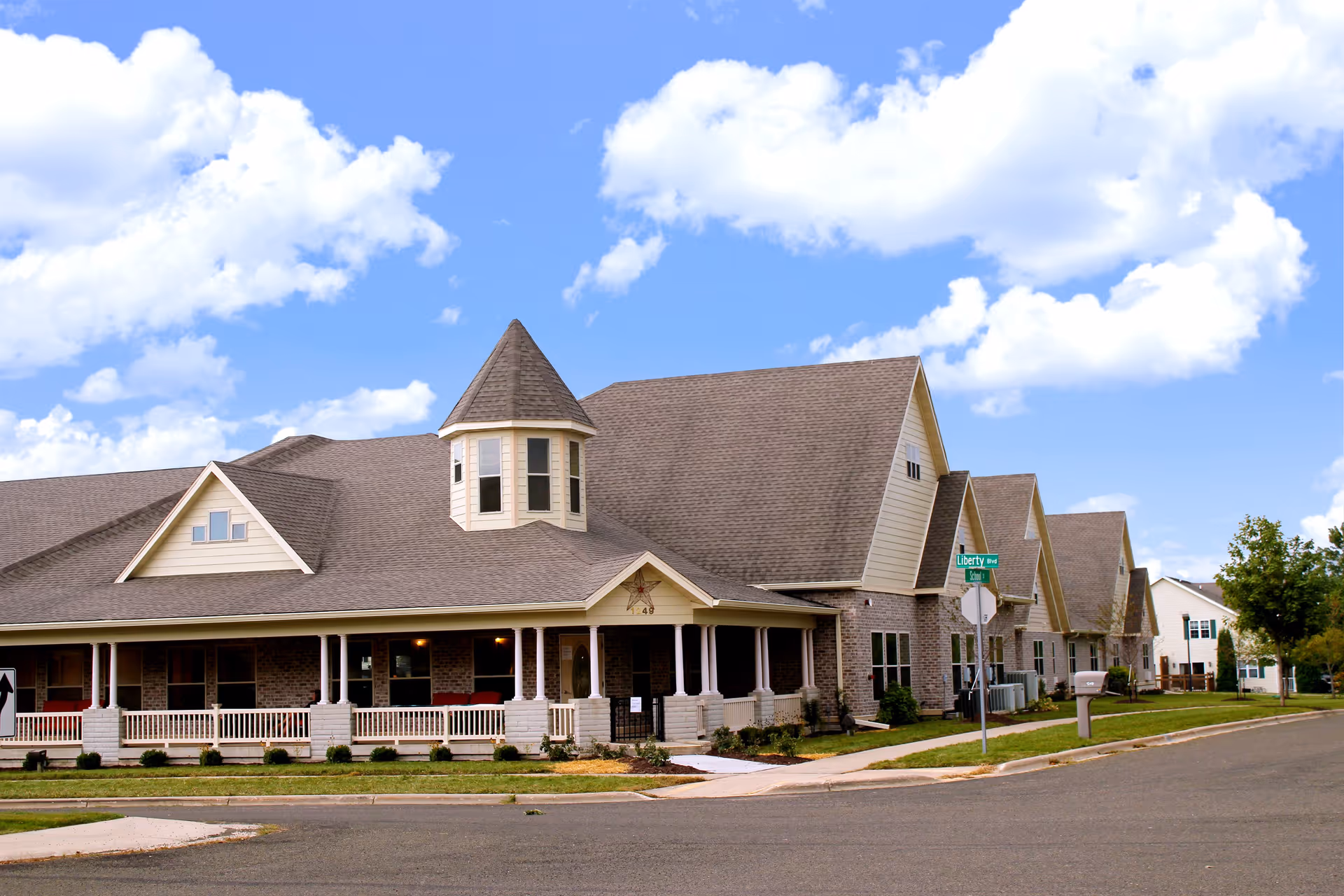 Exterior front view of a large memory-care residential building with a wraparound porch and a small turret under a blue sky.