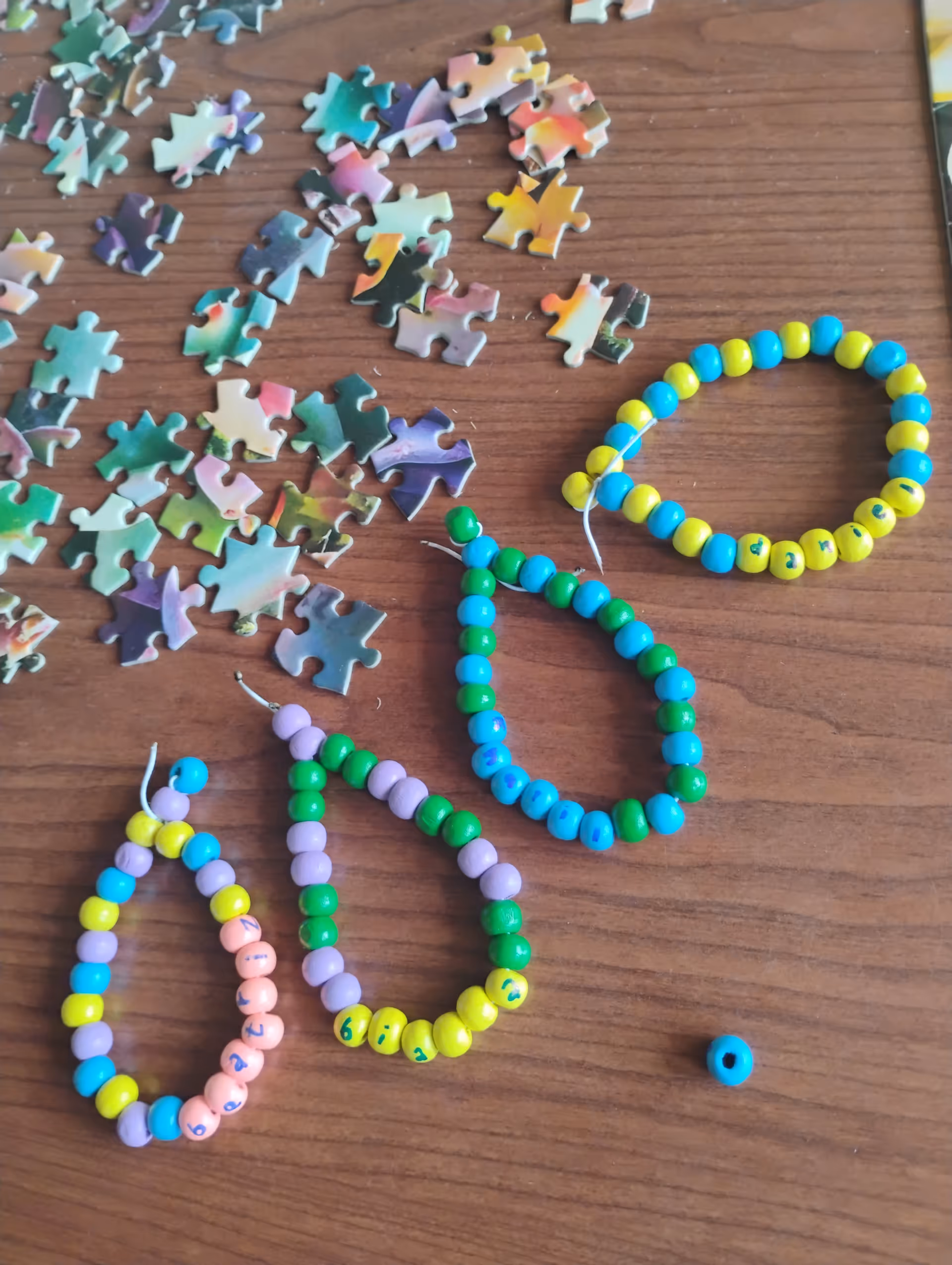 Colorful bead bracelets and scattered puzzle pieces on a wooden table indoors.