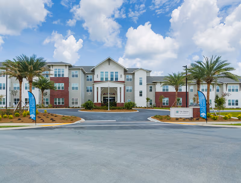 Three-story senior living building front with a covered central entrance, palm trees, and a circular driveway under a blue sky.