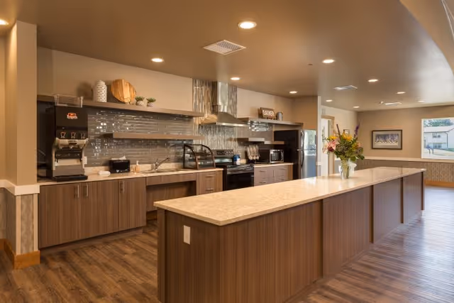 Communal modern kitchen with a long central island, stainless steel appliances, coffee station, and a vase of flowers.