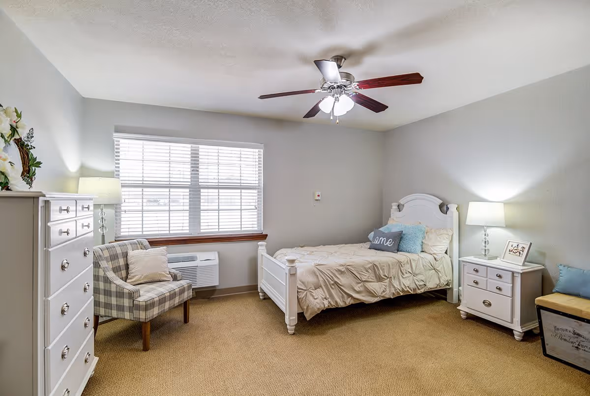 A cozy bedroom with a single white bed adorned with beige bedding and decorative pillows, including one that says 'home'. The room features a white chest of drawers with a lamp and flowers on top, a plaid armchair with a cushion, a white nightstand with a lamp and framed photo, a ceiling fan with lights, and a window with blinds letting in natural light. The walls are painted light gray and the floor is carpeted in beige.