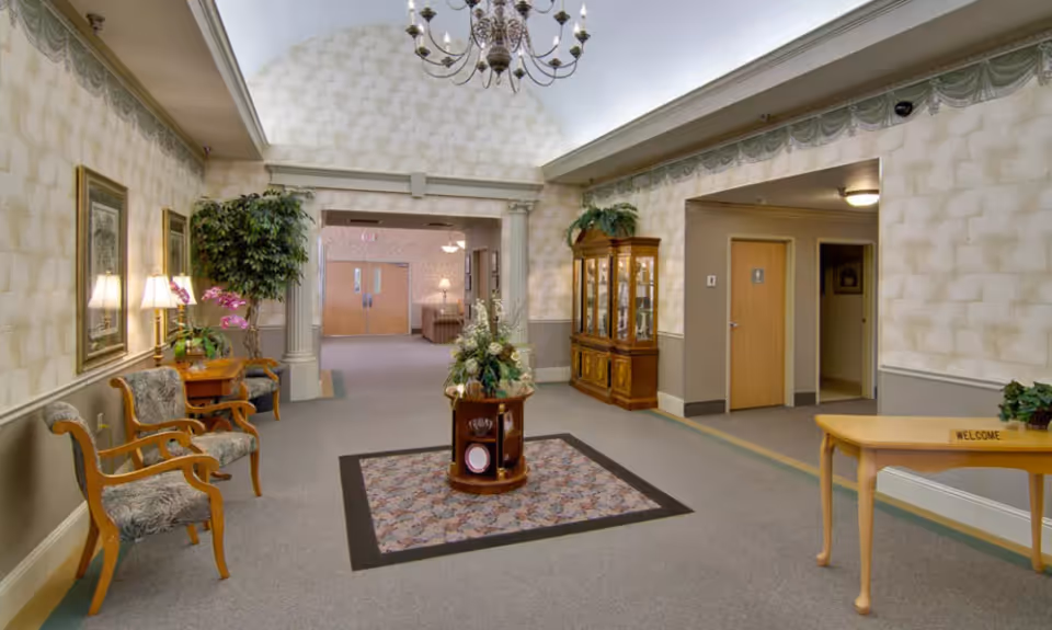 A spacious, well-lit interior hallway of a senior living facility with patterned wallpaper and carpeted floor. There are two wooden chairs with patterned cushions and a small table with lamps and plants on the left side. A round table with a floral arrangement is centered on a decorative rug. On the right side, there is a wooden display cabinet with glass doors and a wooden table with a small 'WELCOME' sign. The hallway leads to double doors and another seating area in the background.