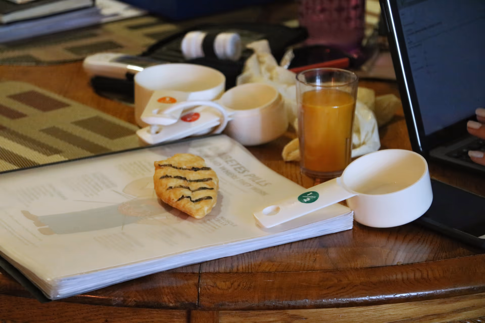 A cluttered wooden table with a pastry, glass of juice, cookbook, and a tablet.