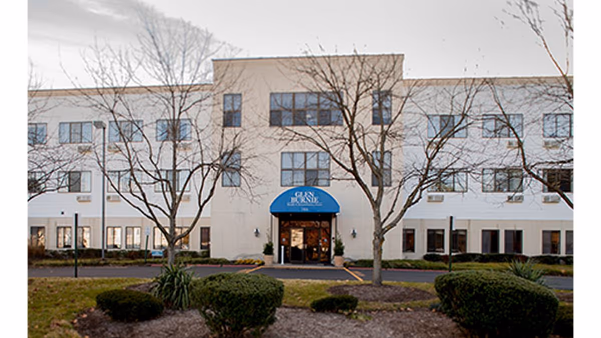 Front exterior view of a three-story senior living facility building with a blue awning above the main entrance that reads 'Glen Burnie'. The building is surrounded by leafless trees and trimmed bushes in a landscaped area.