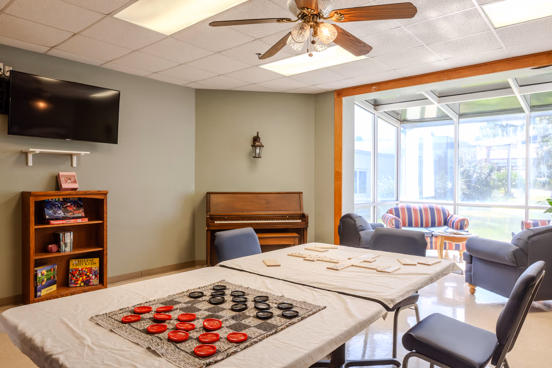 Activity room with tables, chairs, a piano, bookshelves, game pieces, and a glass-walled seating area.