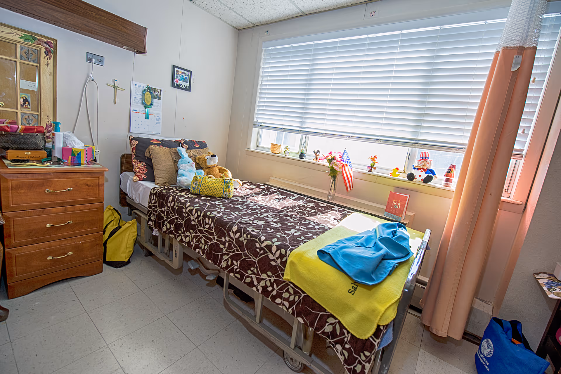 A single bed in a senior living facility room with a brown floral blanket, several pillows, and stuffed animals on it. Next to the bed is a wooden dresser with various personal items on top. A window with white blinds and a windowsill decorated with small figurines, flowers, and an American flag is on the right side. The room has light-colored walls and a tiled floor.