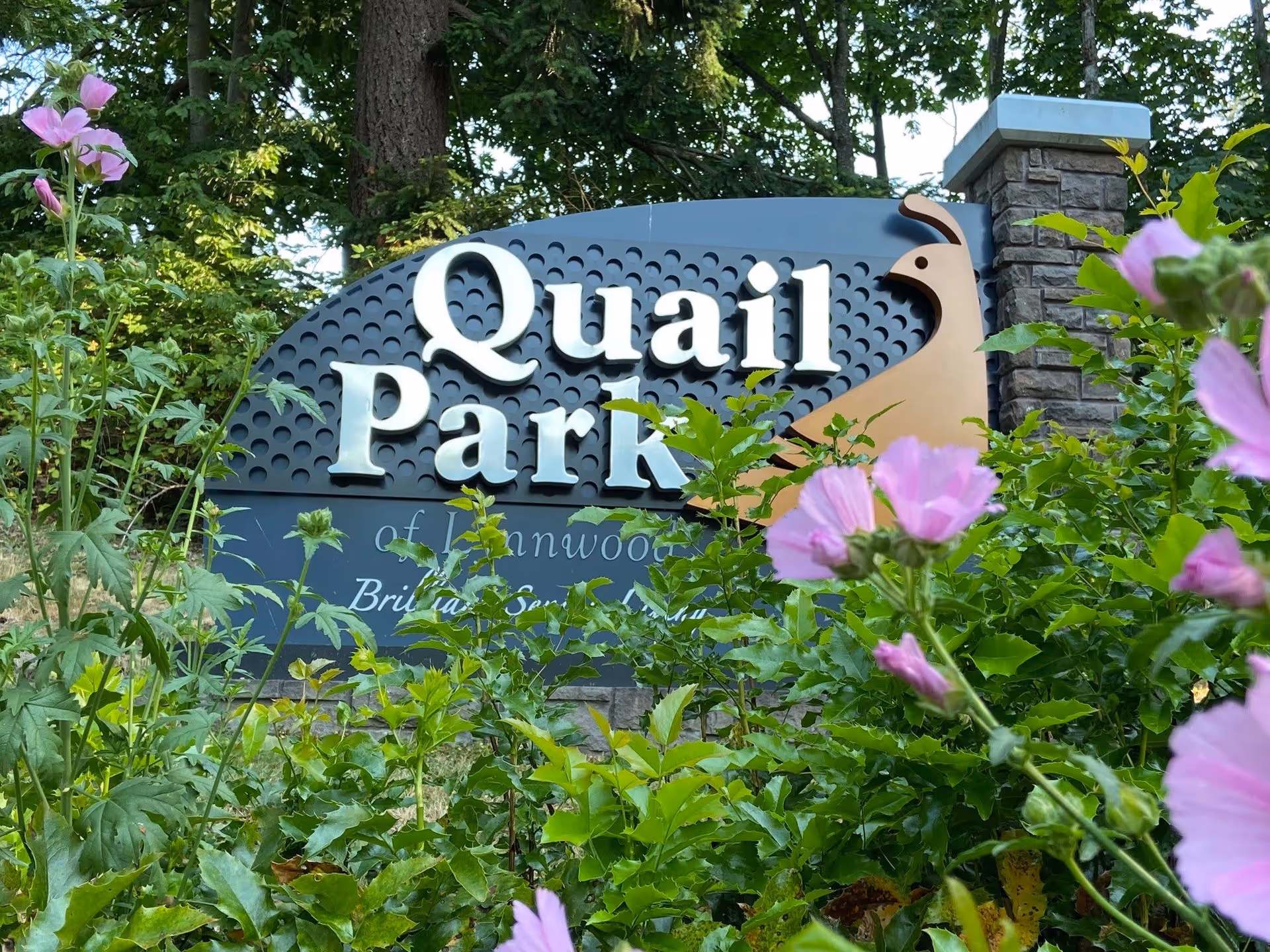 Entrance sign reading "Quail Park" surrounded by green foliage and pink flowers.