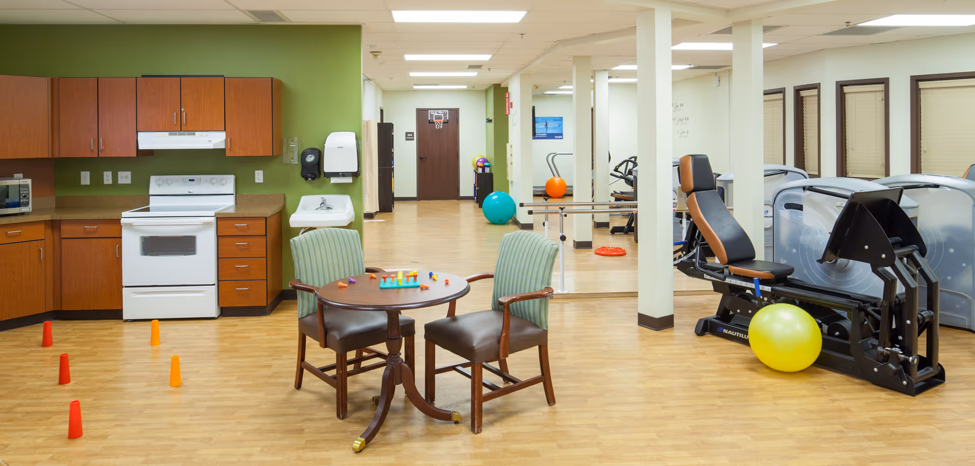 A multi-purpose room in a senior living facility featuring a kitchen area with wooden cabinets, a white stove, and a microwave. In the foreground, there is a small round table with two green cushioned chairs and colorful game pieces on the table. The room also includes exercise equipment such as a leg press machine and stationary bikes, with colorful exercise balls scattered around. The floor is wood, and the walls are painted green and white.