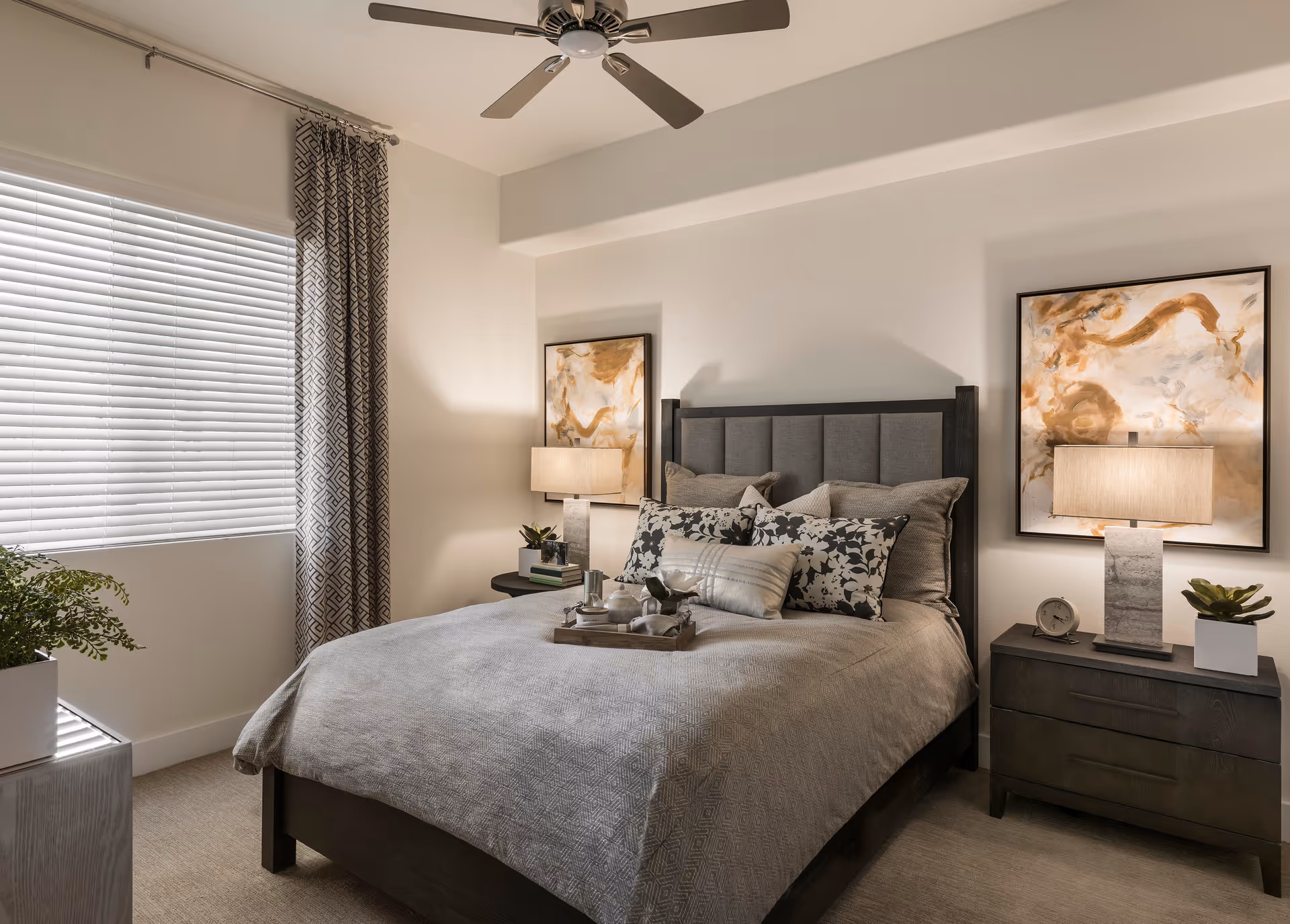 A neatly made bed with a gray upholstered headboard and multiple decorative pillows in a well-lit bedroom. There are two nightstands on either side of the bed, each with a lamp and decorative items. Two abstract paintings hang on the wall above the nightstands. A window with blinds and patterned curtains is on the left side of the room, and a ceiling fan is mounted above.