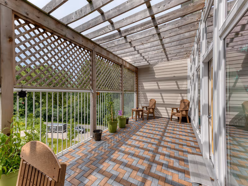 A covered outdoor patio area with a wooden lattice roof and railing. The floor is paved with interlocking bricks in a herringbone pattern. There are several potted plants along the railing and two wooden chairs with a small table between them at the far end. The patio overlooks a green area with trees and a parking lot visible in the background.