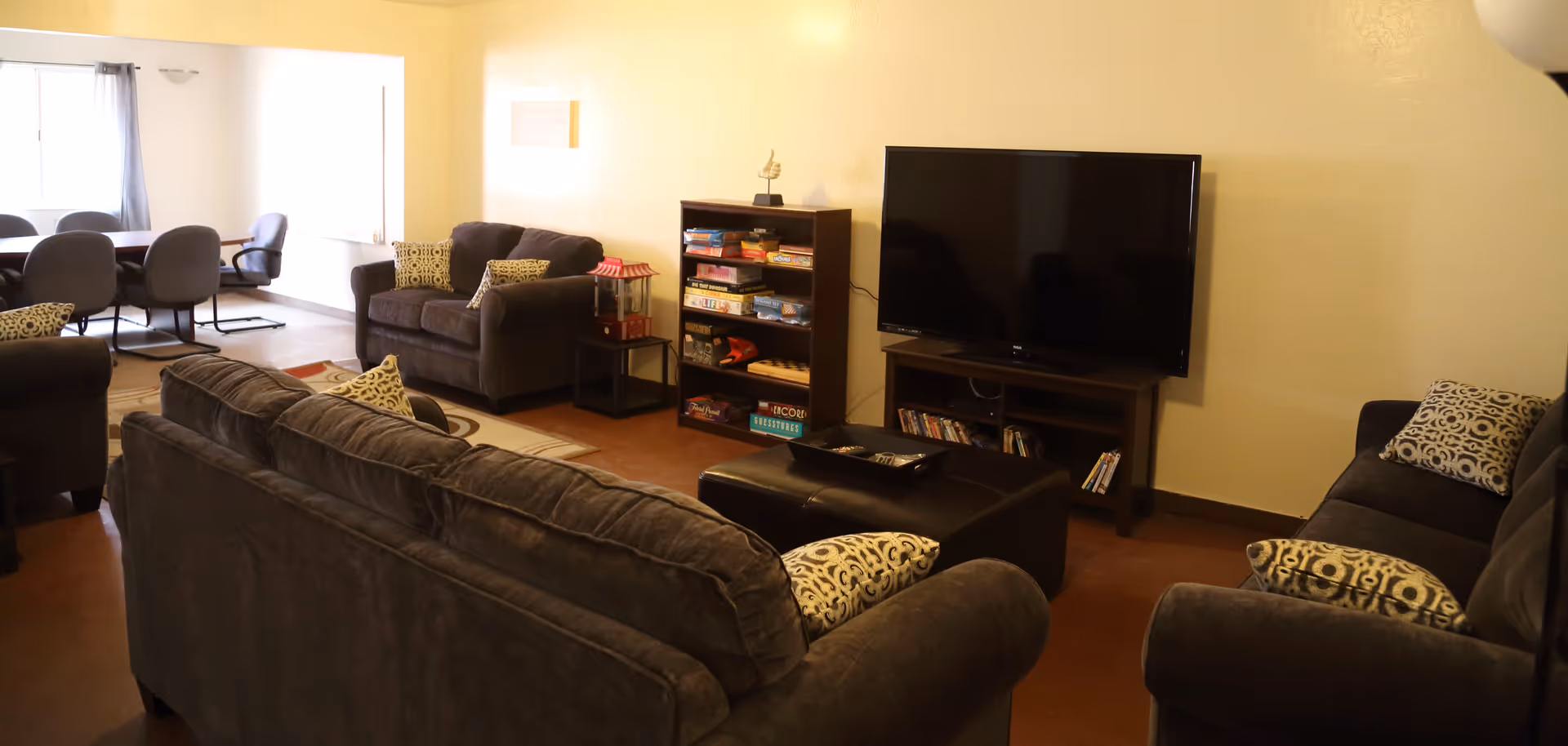 Community lounge with multiple gray sofas, a large flat-screen TV on a stand, shelving with board games, and a dining table visible in the background.